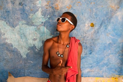 Photo of a young man with no shirt and sunglasses standing in front of a blue wall, Santa Cruz del Islote, Colombia, December 2017, Reinder Nijhoff